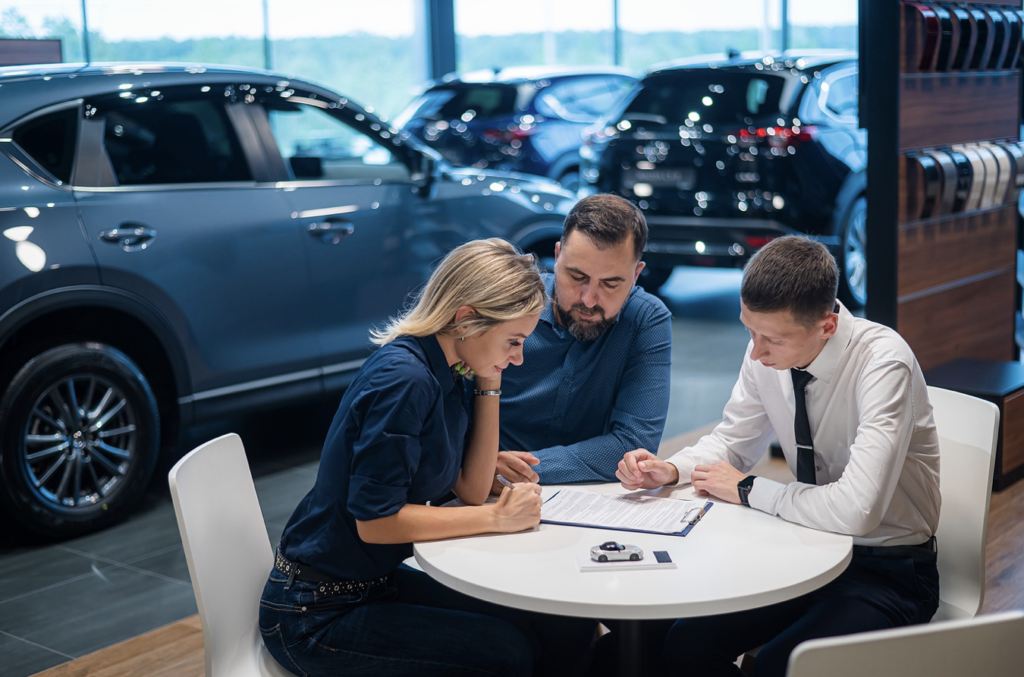 A man and woman clients sitting at a round table at a dealership with the sales person, signing agreements.