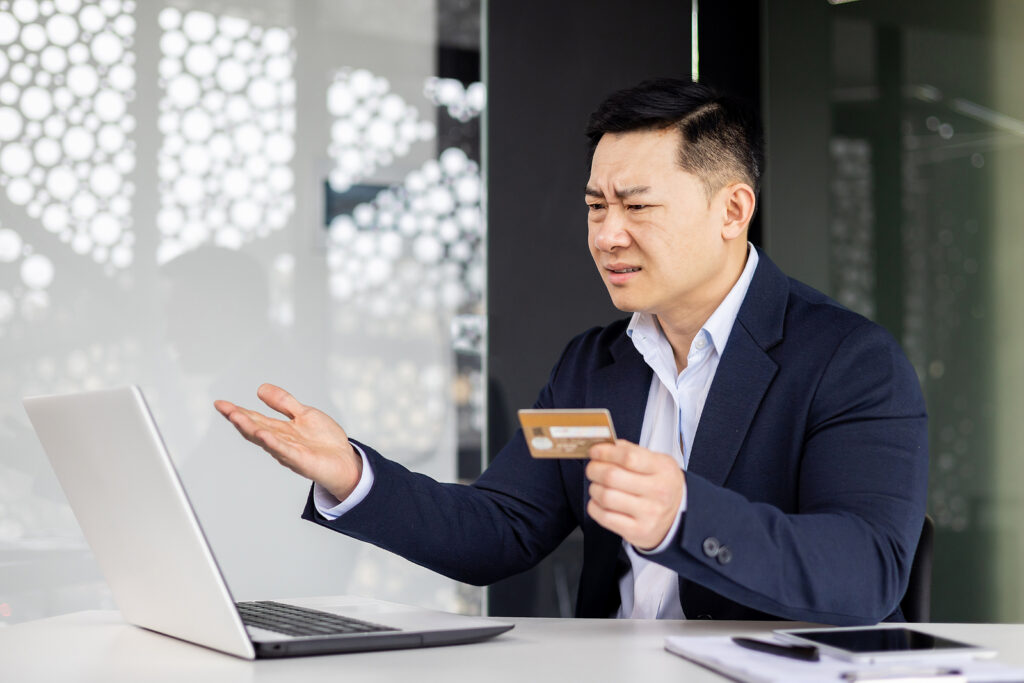 frustrated again business man holding a credit card and his other hand out in confusion looking at a laptop